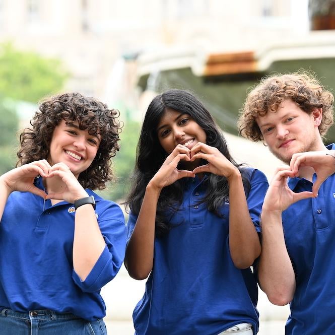 Three students make heart shapes with their hands in front of a fountain